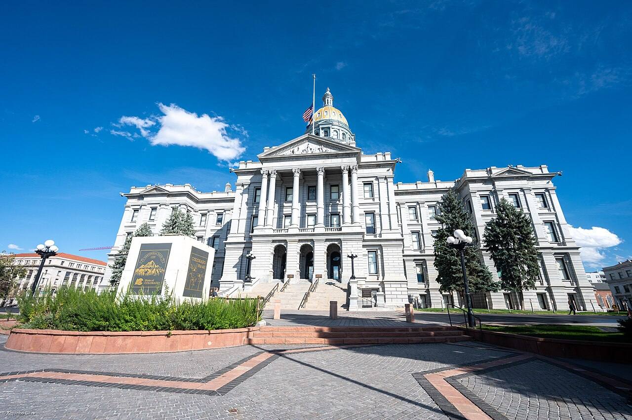 Fachada principal del Capitolio del Estado de Colorado en Denver, con su cúpula dorada, el pórtico de columnas y un monumento con el rótulo "COLORADO" en el jardín delantero, bajo cielo despejado.
