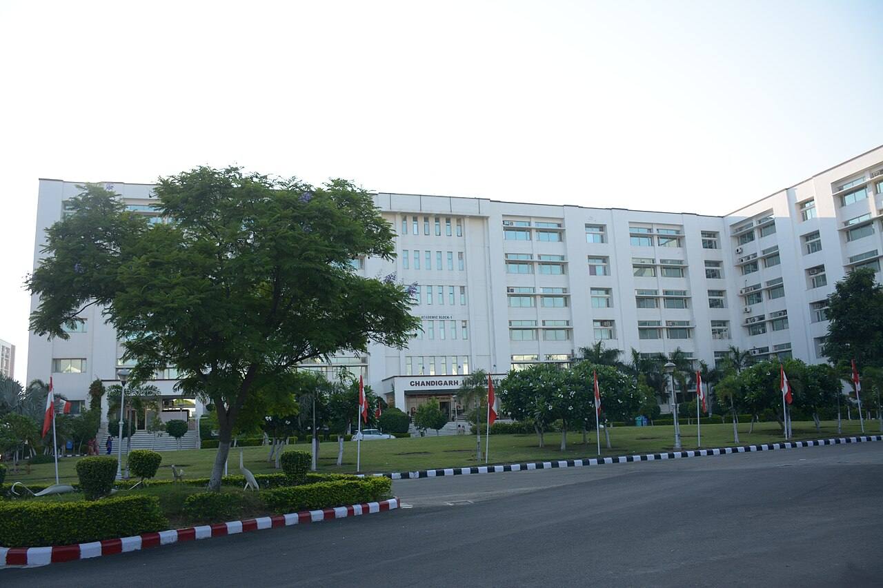 Fachada del Bloque 1 de la Universidad de Chandigarh, con el rotulo "CHANDIGARH UNIVERSITY" sobre la entrada, cesped, banderas y coches aparcados delante del edificio