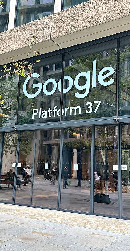 Rótulo iluminado «Google Platform 37» sobre la fachada acristalada de la entrada principal del campus de Google en King's Cross, Londres, con transeúntes junto a las puertas correderas