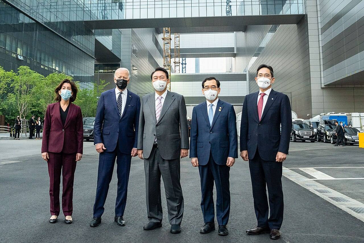 El presidente de EE. UU. Joe Biden junto al presidente surcoreano Yoon Suk-yeol y otros funcionarios frente al campus de fabricación de semiconductores de Samsung Electronics en Pyeongtaek, Corea del Sur, con un edificio en construcción de fondo (mayo de 2022)