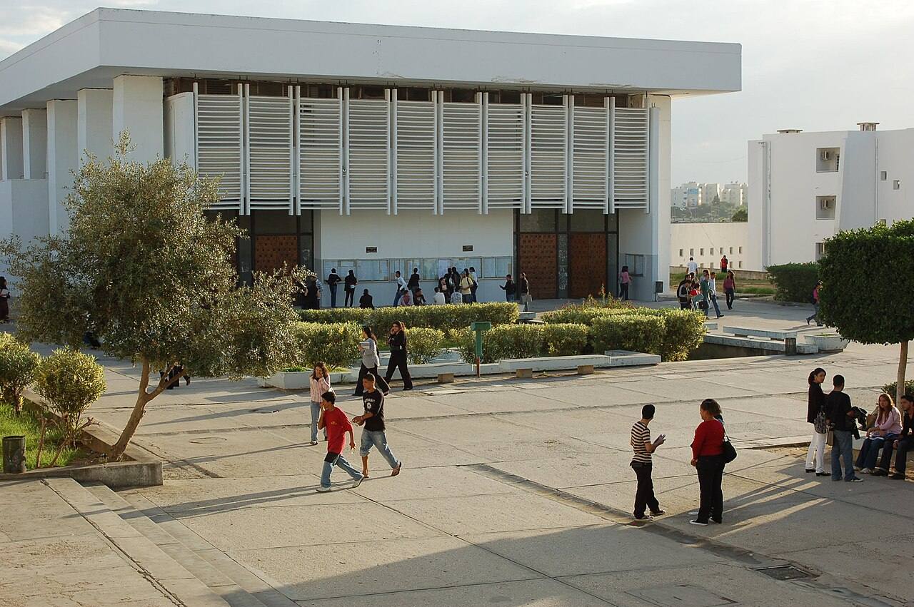 Edificio de fachada blanca con brise-soleil en el campus de la Universidad de Túnez El Manar, con estudiantes cruzando la explanada exterior