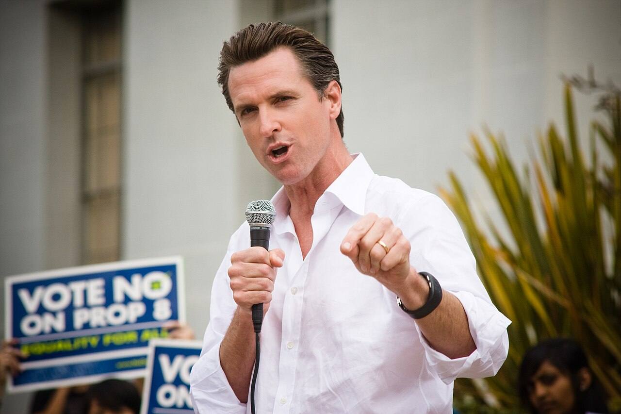 El gobernador de California, Gavin Newsom, hablando en un podio con la bandera de Estados Unidos de fondo.
