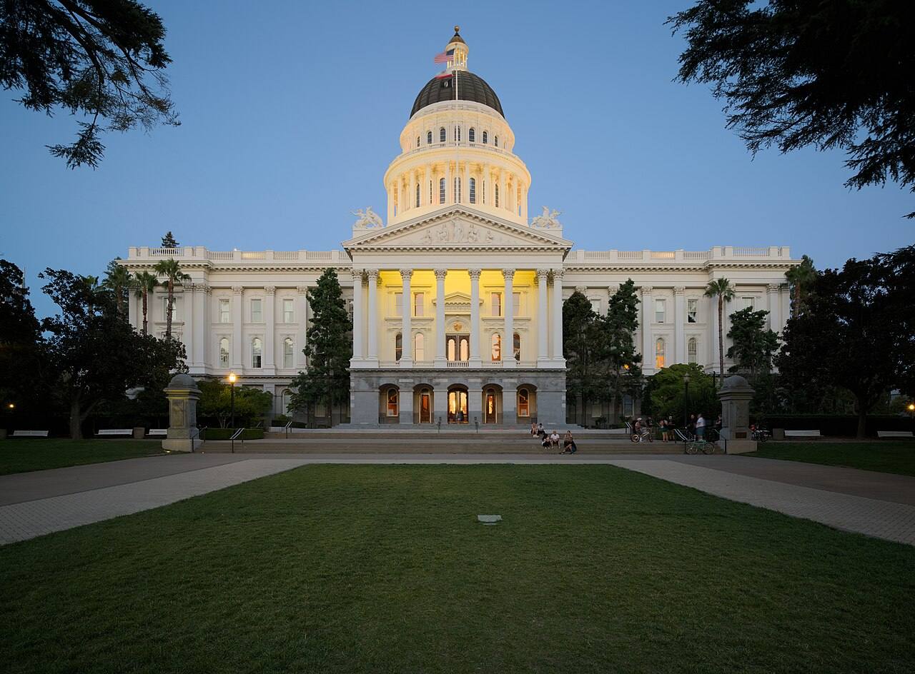 Fachada frontal del Capitolio del Estado de California iluminada al anochecer, con cúpula dorada y columnata clásica bajo un cielo azul oscuro.