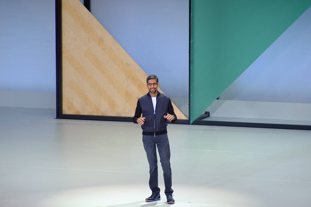 Sundar Pichai, consejero delegado de Google, solo en el centro del escenario del congreso de desarrolladores Google I/O de 2017: chaqueta azul marino con cremallera sobre camiseta blanca, hablando con las manos abiertas, sobre un suelo blanco y ante un decorado de grandes bloques de color —beis, verde y azul— que ocupa todo el fondo. Fotografiado el 17 de mayo de 2017.