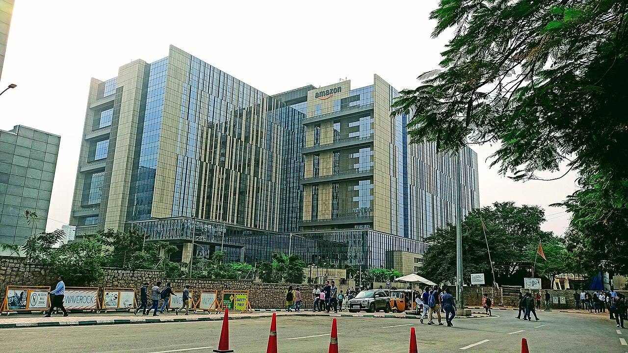 Fachada del campus de Amazon en el distrito financiero de Hyderabad, India, con el logotipo "amazon" en el edificio, un auto-rickshaw y peatones en la calle en primer plano