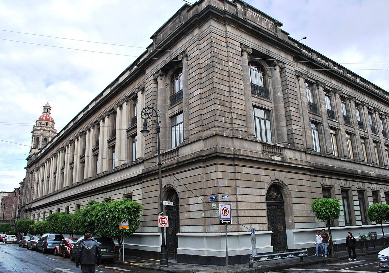 Esquina del edificio histórico de la Secretaría de Educación Pública (SEP) en el Centro Histórico de la Ciudad de México, con la torre de un antiguo convento al fondo y peatones en la acera.