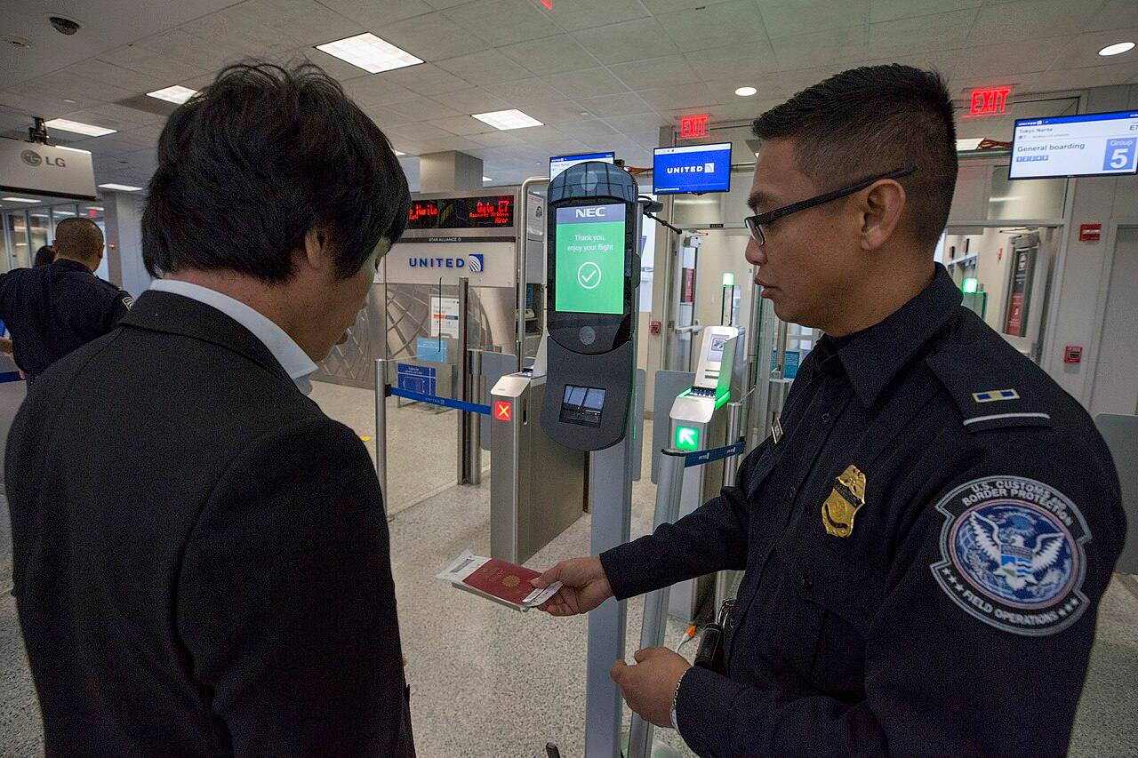Un oficial de la Oficina de Campo de la Aduanas y Protección Fronteriza de EE. UU. (CBP), con el parche "U.S. Customs and Border Protection / Field Operations" visible en el uniforme, compara el pasaporte de un pasajero con la foto captada por un quiosco NEC de reconocimiento facial biométrico, junto a la puerta de embarque del aeropuerto de Houston