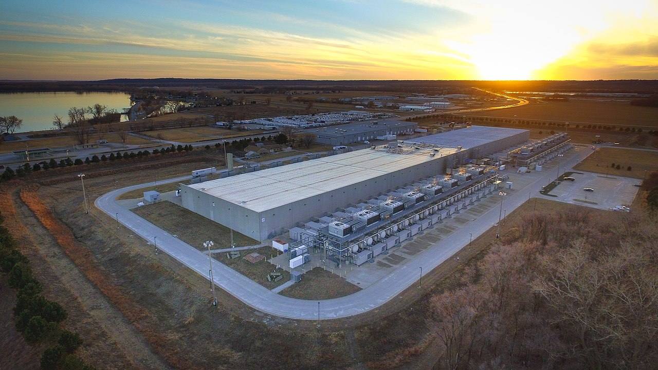Vista aérea al atardecer de un centro de datos de Google en Council Bluffs, Iowa: una gran nave industrial con hileras de equipos de refrigeración a lo largo del lateral, aparcamientos y viales de acceso alrededor.
