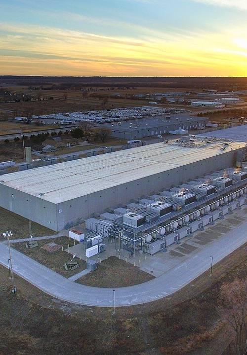 Vista aérea al atardecer de un centro de datos de Google en Council Bluffs, Iowa: una gran nave industrial con hileras de equipos de refrigeración a lo largo del lateral, aparcamientos y viales de acceso alrededor.