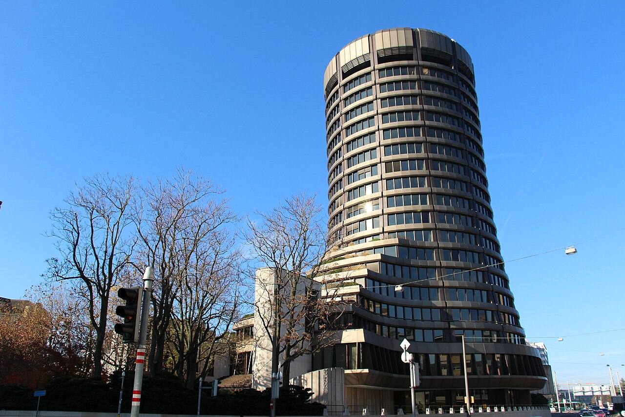 Torre cilíndrica de hormigón de la sede del Banco de Pagos Internacionales en Basilea, vista desde la calle bajo cielo despejado, con árboles sin hojas en primer plano