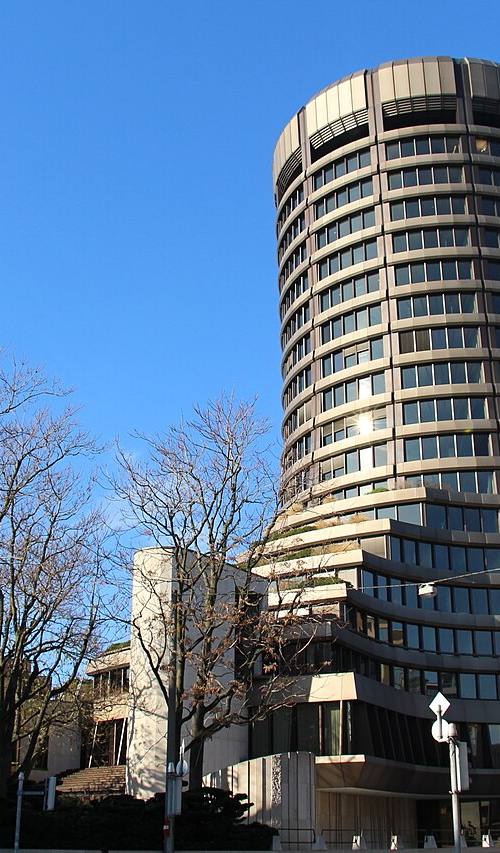 Torre cilíndrica de hormigón de la sede del Banco de Pagos Internacionales en Basilea, vista desde la calle bajo cielo despejado, con árboles sin hojas en primer plano