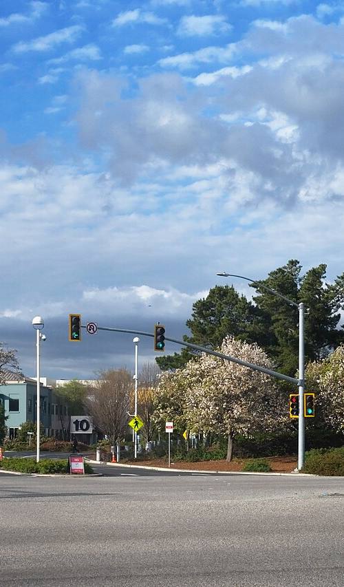 Entrada al campus de Meta Platforms en Menlo Park, California: cruce de calles arbolado con un cartel iluminado que dice "Meta — 1 Hacker Way" junto a semáforos y edificios bajos de oficinas al fondo