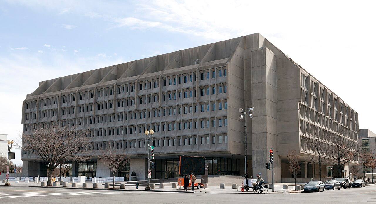 Vista en ángulo de la fachada de hormigón con ventanas en zigzag del edificio Hubert H. Humphrey, sede del Departamento de Salud y Servicios Humanos de EE. UU. en Washington D.C., con calle y aceras en primer plano bajo cielo despejado.