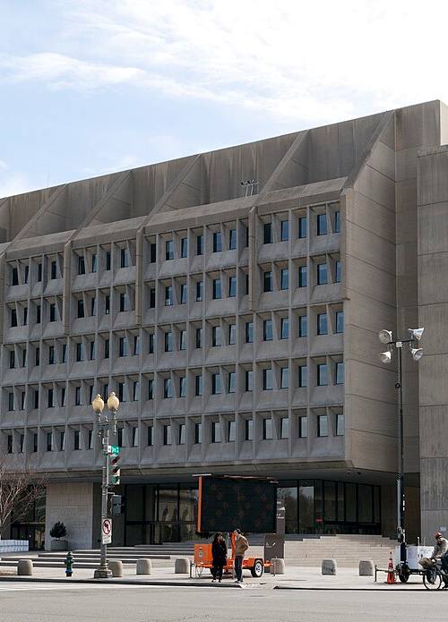 Vista en ángulo de la fachada de hormigón con ventanas en zigzag del edificio Hubert H. Humphrey, sede del Departamento de Salud y Servicios Humanos de EE. UU. en Washington D.C., con calle y aceras en primer plano bajo cielo despejado.