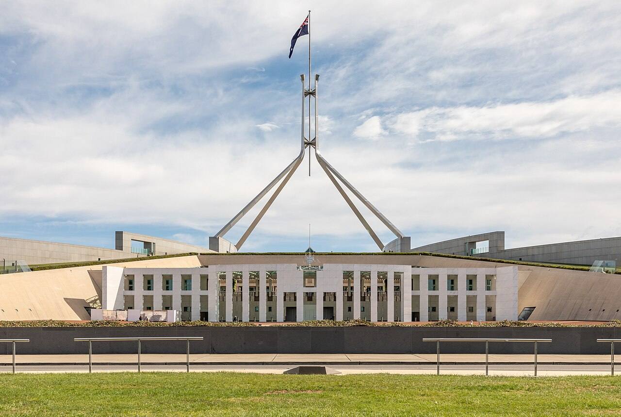 Fachada simétrica del Parlamento de Australia en Canberra, con el mástil y la bandera nacional sobre el tejado, bajo un cielo despejado con nubes.