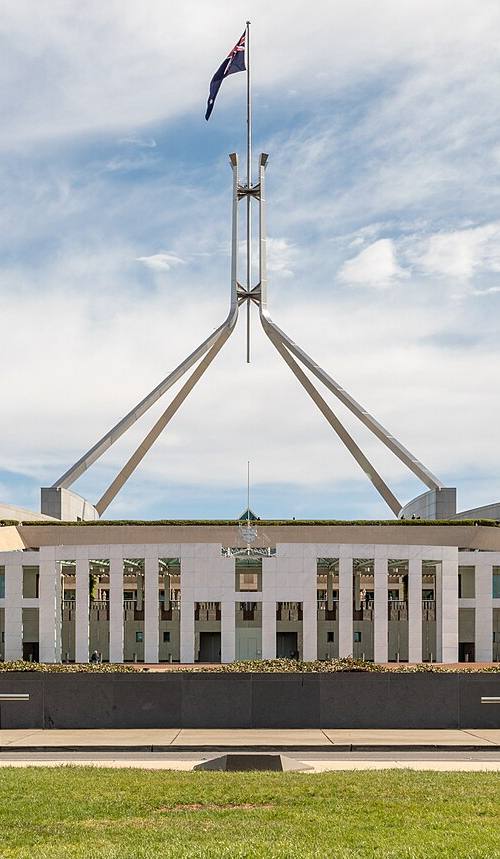 Fachada simétrica del Parlamento de Australia en Canberra, con el mástil y la bandera nacional sobre el tejado, bajo un cielo despejado con nubes.