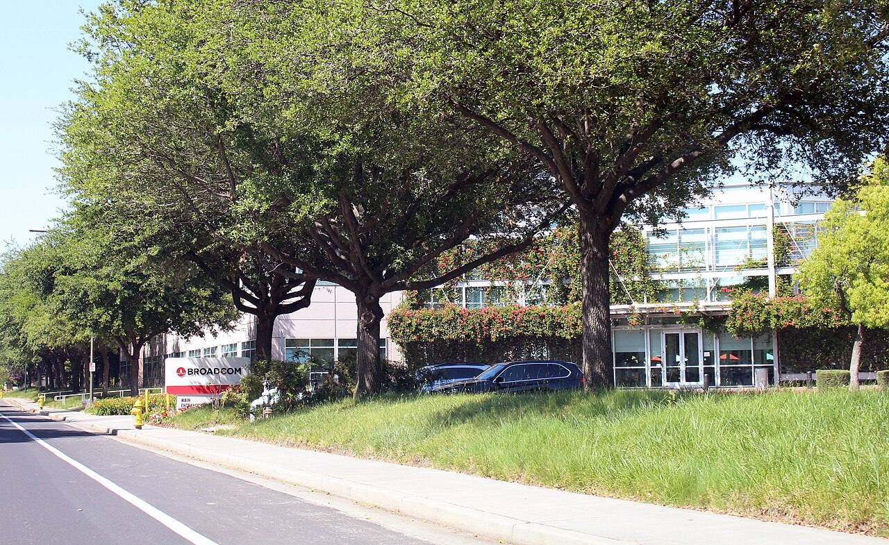 Letrero corporativo de Broadcom junto a la entrada principal de un edificio de oficinas de su sede en San José, California, rodeado de árboles y jardines, con coches aparcados frente a la puerta.